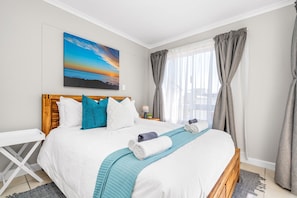 A neatly made bed with white and blue bedding, a wooden headboard, and a view of the sea through large windows with grey curtains. - Coral Grove (Cape Town)