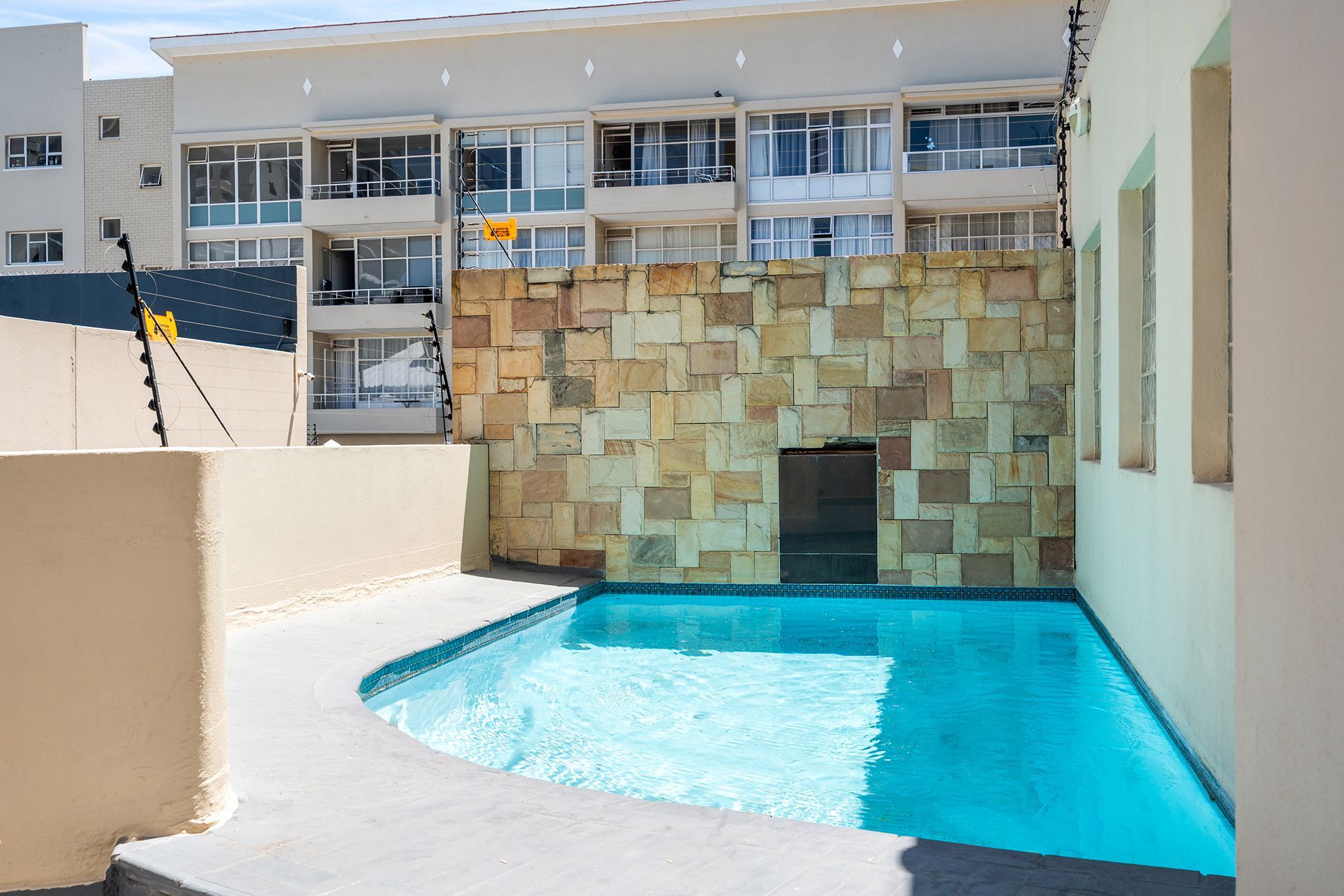 A swimming pool with a stone wall backdrop, adjacent to a multi-story building with balconies.