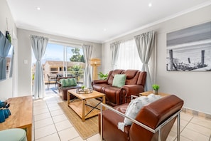 A modern living room with a brown leather sofa, a wooden coffee table, and a dining table with chairs. There is a large window with curtains, a painting on the wall, and a view of a building outside. - Coral Grove (Cape Town)