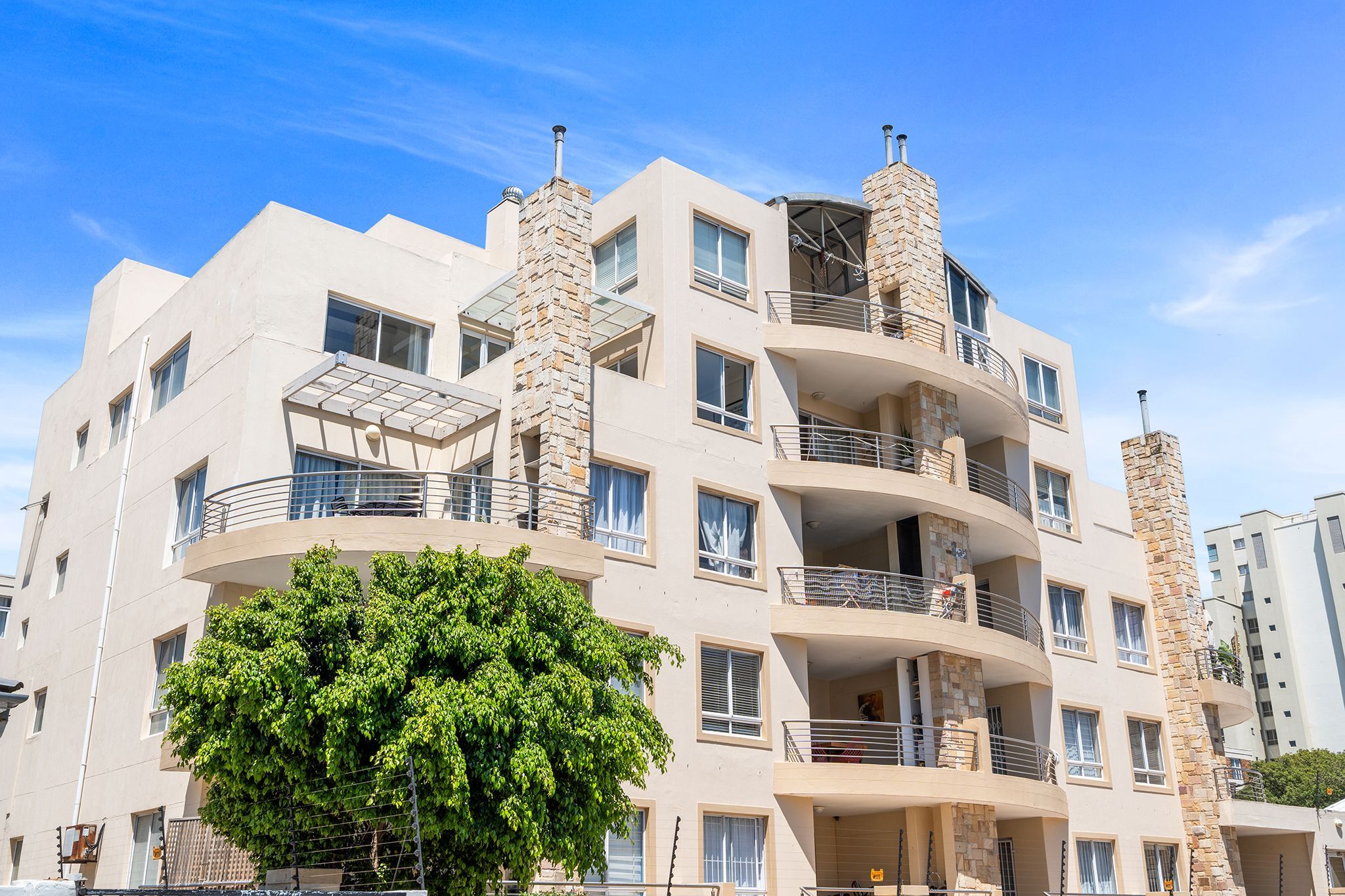 A multi-story residential building with balconies, large windows, and a tree in front.
