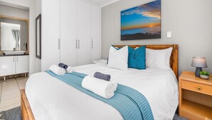 A neatly made bed with white and blue bedding, a wooden headboard, and a nightstand with a lamp and a small plant. - Coral Grove (Cape Town)