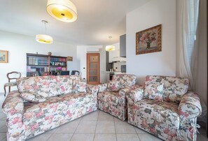 A living room with a floral sofa, a bookshelf, and a dining area with chairs. - The Terrace on the Gulf of Alghero 1 (Alghero)