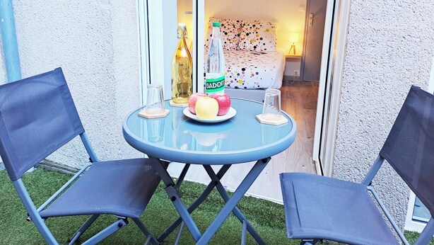Una pequeña zona de estar al aire libre con una mesa azul y dos sillas, una botella de vino y un plato con manzanas.