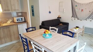 Una cocina moderna con microondas, pileta y muebles de madera. Hay una mesa de comedor con sillas y un cuenco con fruta. Un sofá negro está apoyado en la pared y una pequeña mesa con una planta en la esquina.