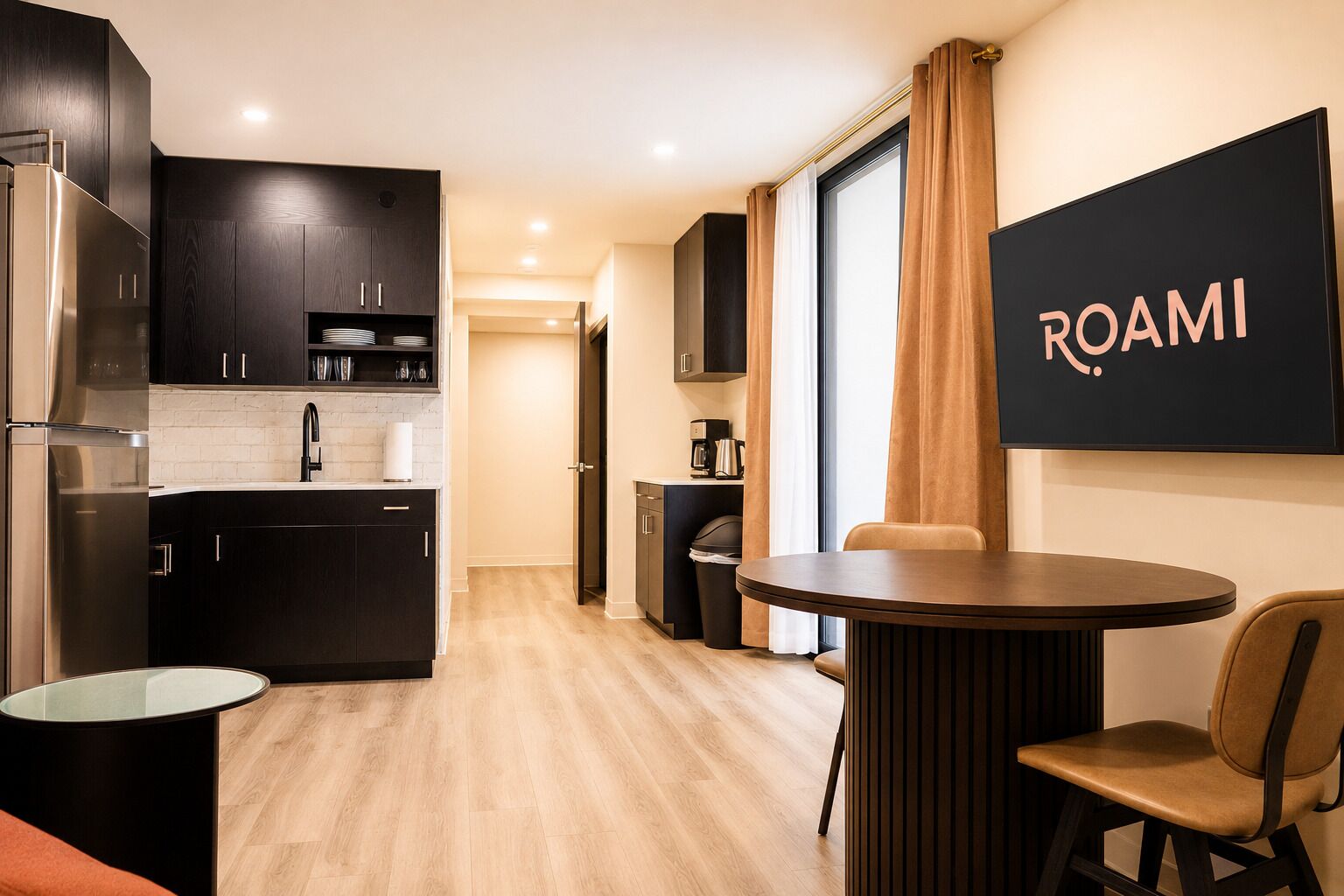 Una cucina moderna con mobili scuri, un frigorifero in acciaio inossidabile, un tavolo rotondo con una sola sedia e un cartello a parete con su scritto 'ROAMI'.