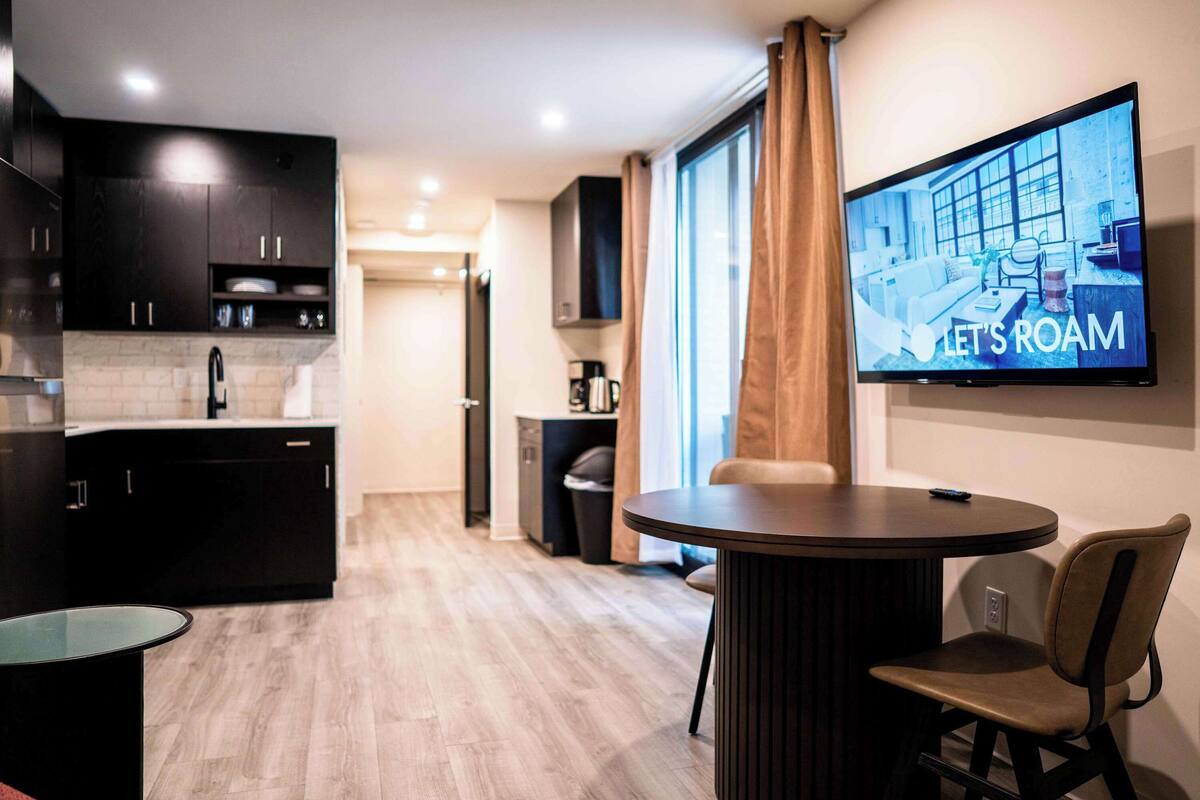 Een moderne keuken met donkere kastjes, een ronde eettafel en een flatscreen televisie waarop 'LET'S ROAM' staat.