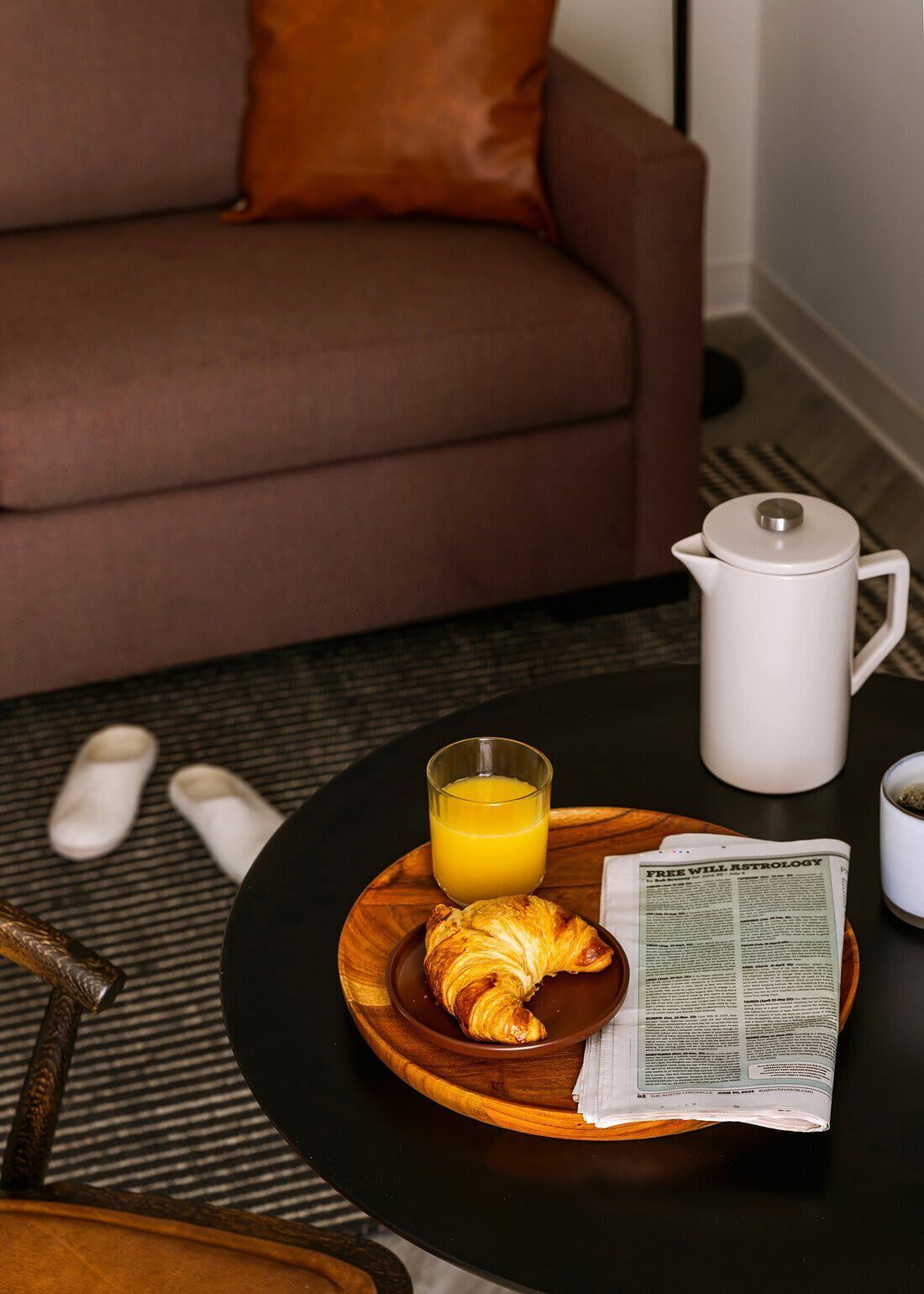 En bricka med frukost bestående av en croissant, apelsinjuice och en tidning, placerad på ett träbord.