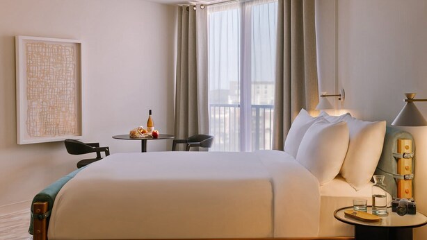 A hotel room with a large bed, a small table with a bottle and fruit, a chair, a lamp, and a framed picture on the wall. - Roami at The Fitz (St. Paul)