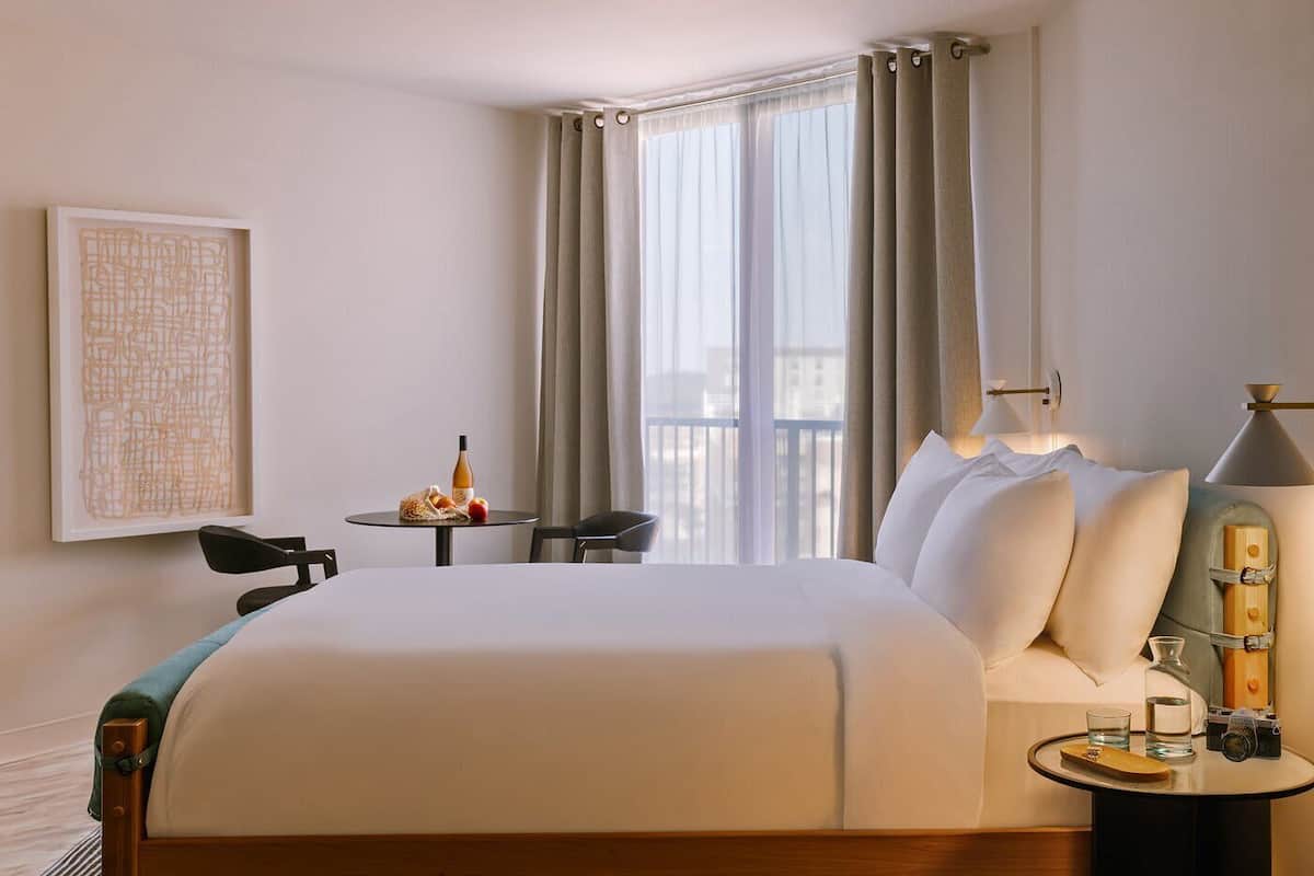 Hotelkamer met een groot bed, een klein tafeltje met een fles en fruit, een stoel, een lamp en een lijstje aan de muur.