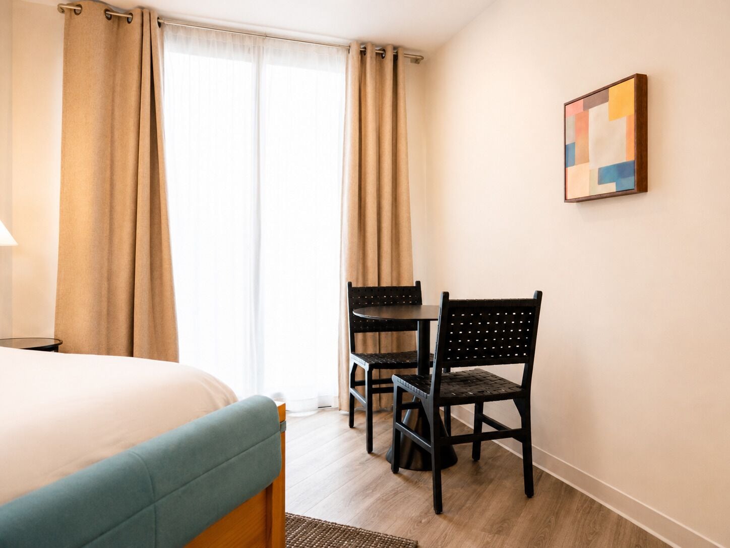 A hotel room with a bed, a desk, two chairs, and a painting on the wall.