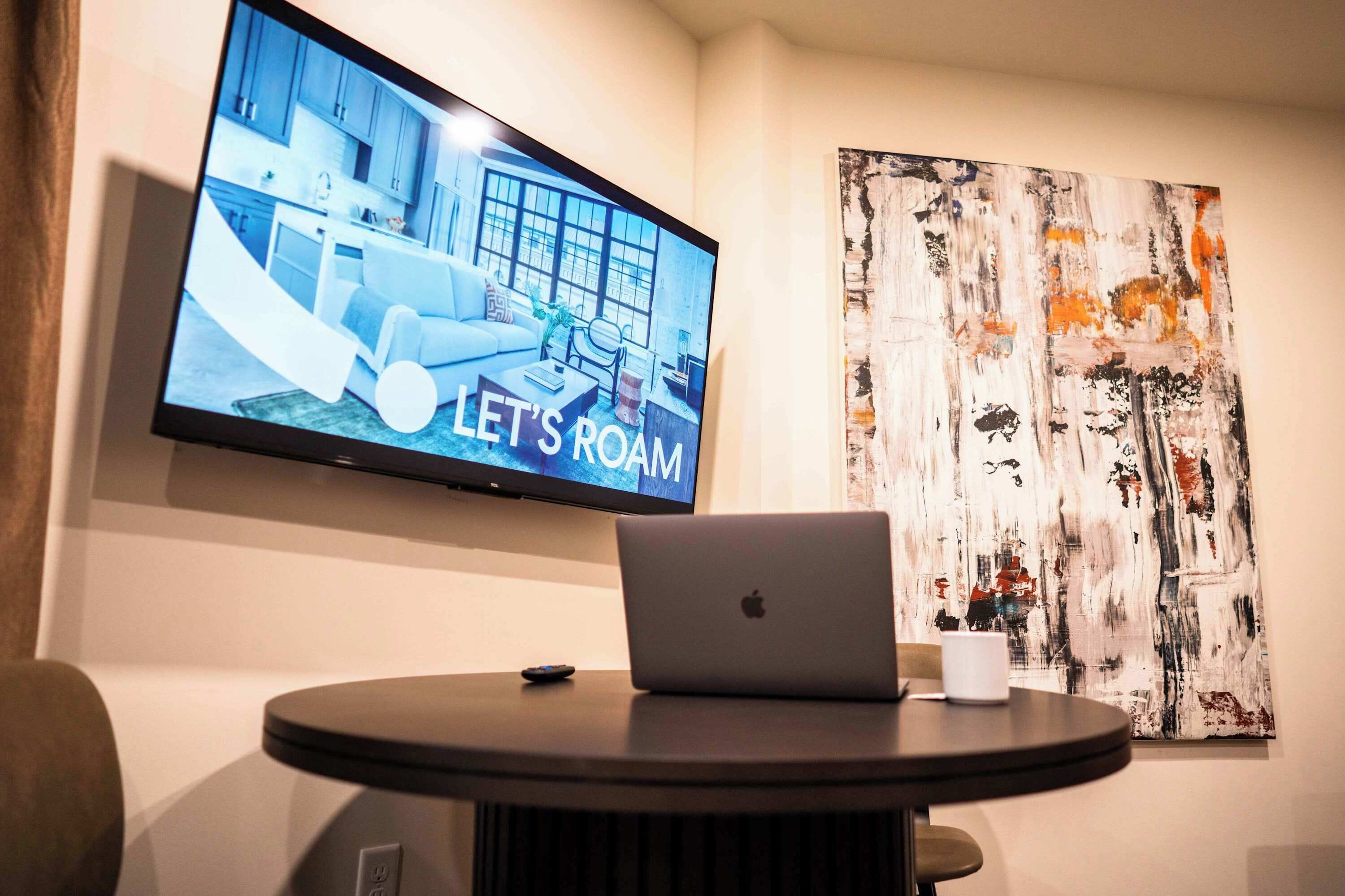 A modern room with a flat-screen TV displaying a room interior, a laptop on a table, and an abstract painting on the wall.