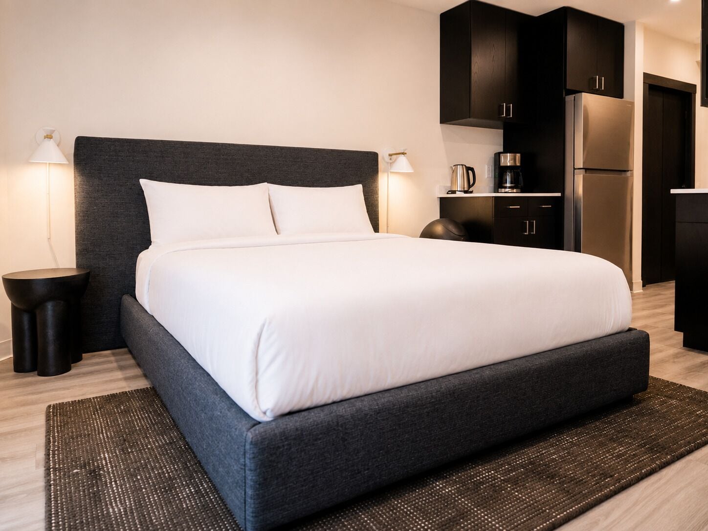 Una camera d'albergo moderna con un letto grande, una testata del letto scura, comodini e un angolo cottura.