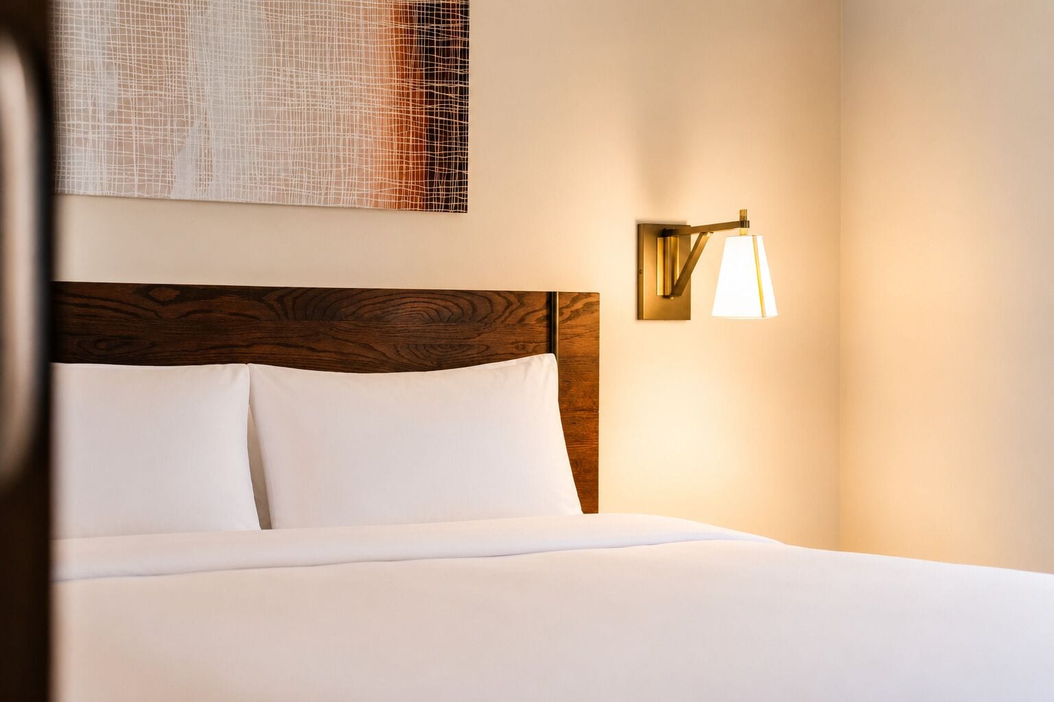 A neatly made bed with white linens, a wooden headboard, and a wall-mounted lamp.
