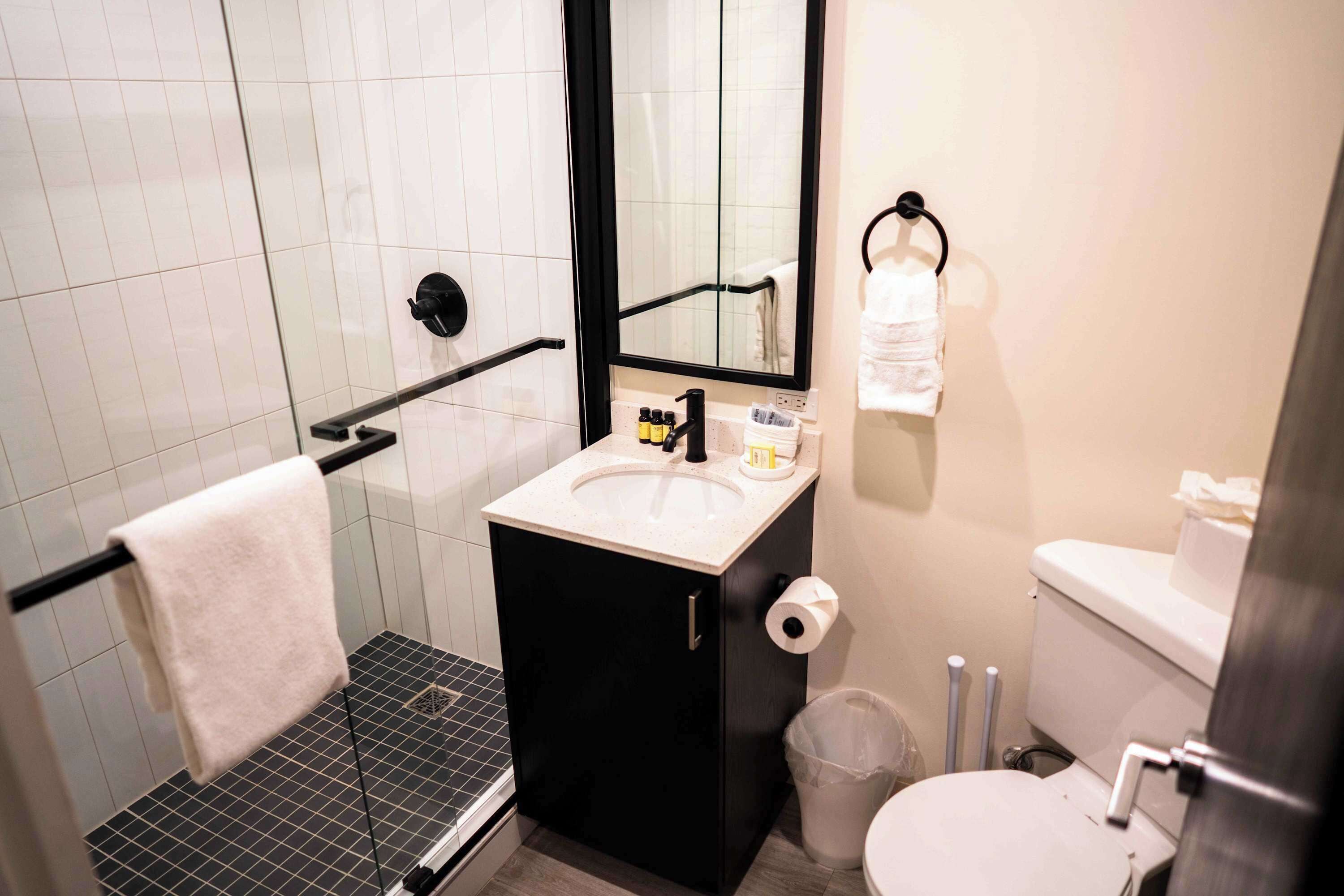 Een badkamer met een douchecabine van glas, een wastafel met spiegel, een toilet en een prullenbak.