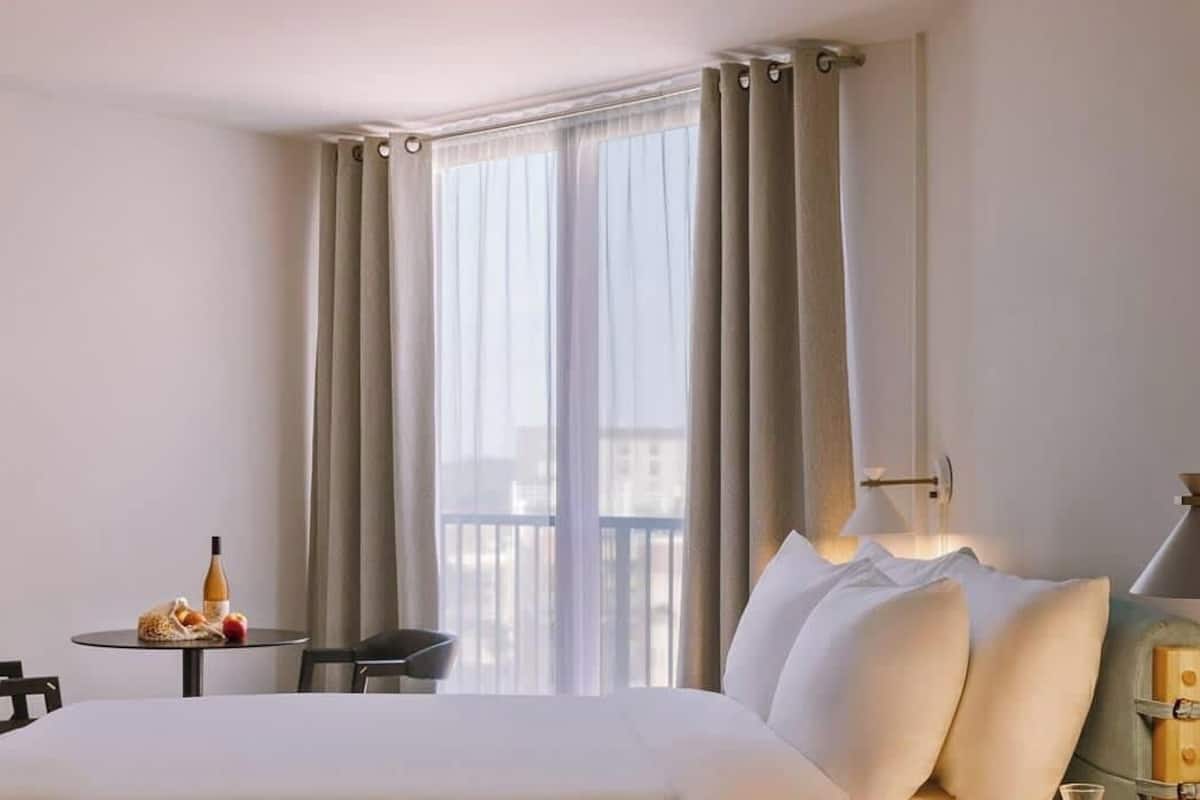 Een hotelkamer met een bed, een klein tafeltje met een fles en fruit, een stoel, een lamp en een raam met gordijnen.