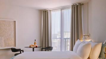 Habitación de hotel con cama, una mesita con una botella y fruta, una silla, una lámpara y un ventanal con cortinas.