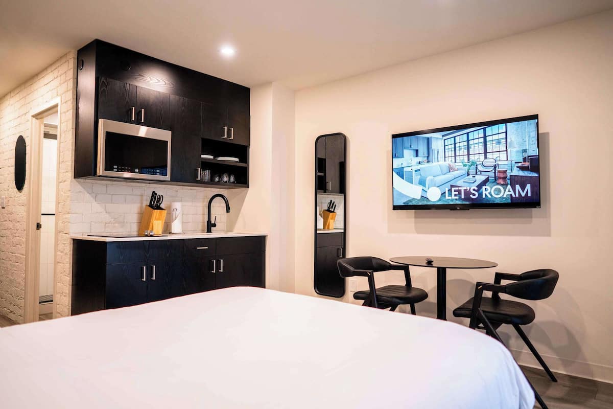 Een moderne hotelkamer met een bed, een televisie waarop een kamer te zien is, een keuken met kasten en een eettafel met stoelen.