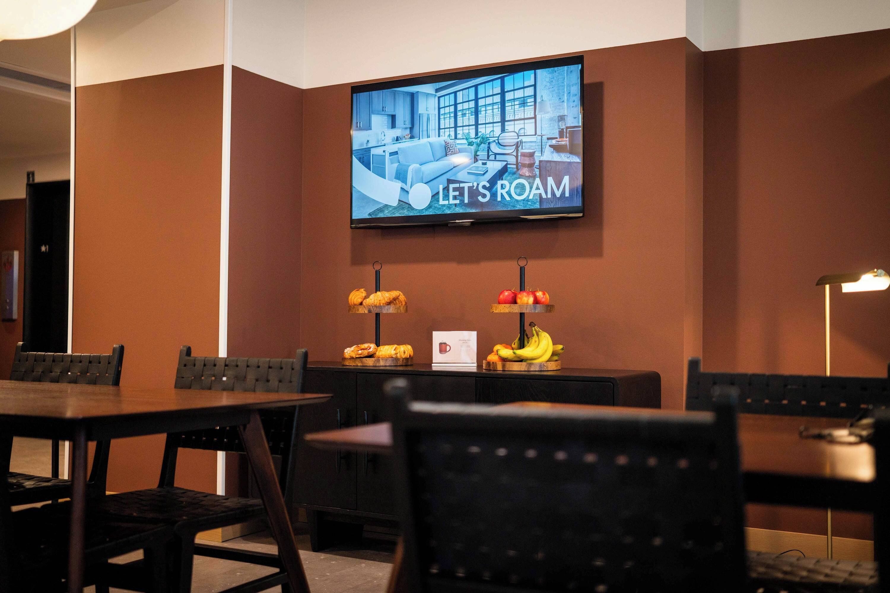 A hotel room with a television displaying 'LET'S ROAM,' a dining area with a table and chairs, and a fruit display.