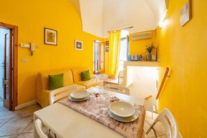 Un comedor con una mesa dispuesta para dos personas, un sofá con cojines y una zona de cocina al fondo.