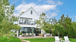 A modern two-story house with a large glass front, a dining area with outdoor furniture, and a well-maintained lawn. - Gorgeous 3-bedroom lakefront house with AC on charming Big Cedar Lake! (West Bend)