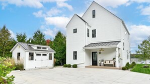 A two-story white house with a garage, a covered porch, and a view of a lake. - Gorgeous 3-bedroom lakefront house with AC on charming Big Cedar Lake! (West Bend)