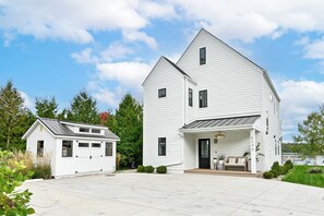 A two-story white house with a garage, a covered porch, and a view of a lake. - Gorgeous 3-bedroom lakefront house with AC on charming Big Cedar Lake! (West Bend)