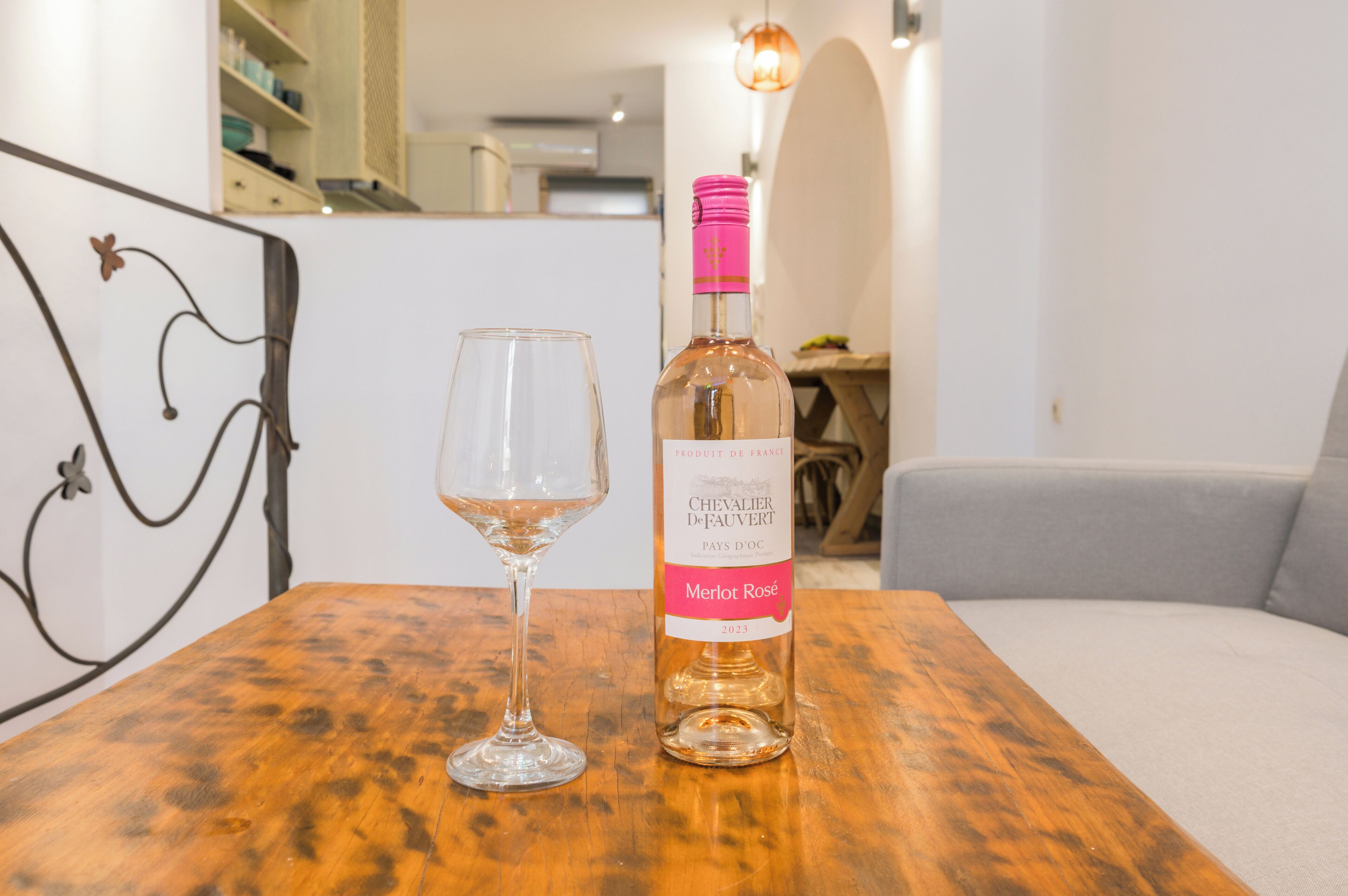 A bottle of Merlot Rosé and a wine glass on a wooden table in a room with a modern design, including a metal art piece and a light fixture.