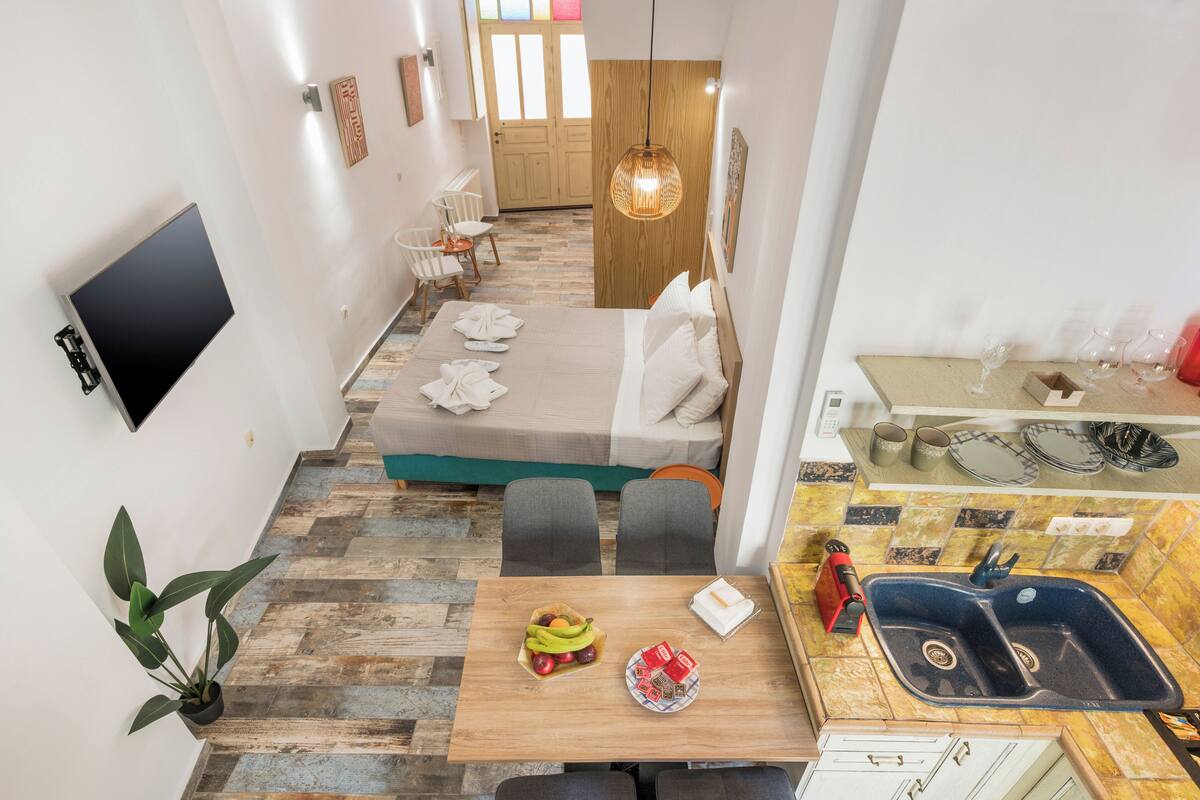 A compact hotel room with a bed, a dining area with a table and chairs, a small kitchen with a sink and a TV mounted on the wall.