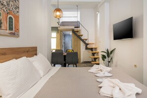 Moderni hotellihuone, jossa on sänky, ruokailutila, portaikko ja seinälle asennettu televisio.