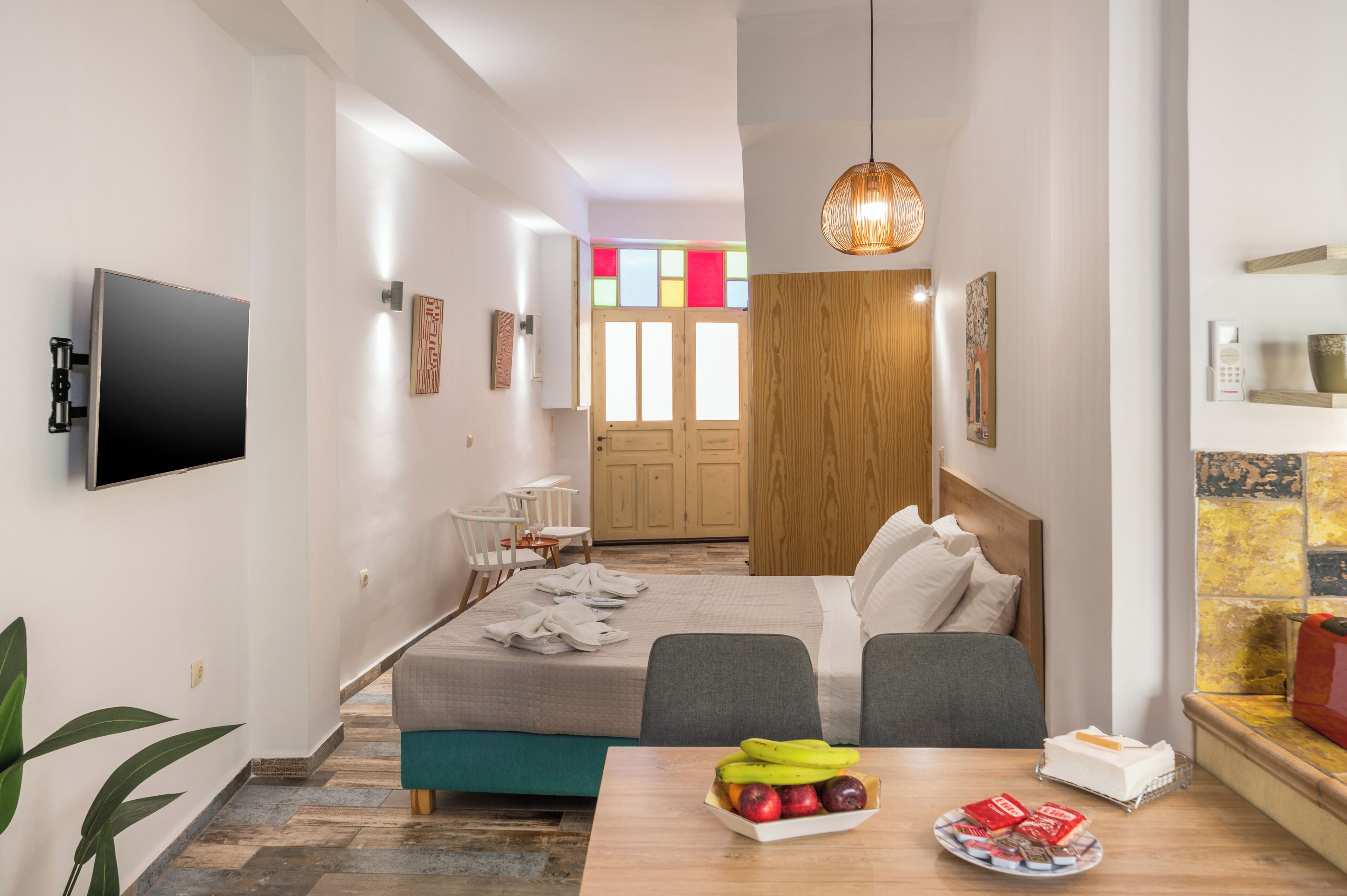 A hotel room with a bed, bedside table with fruit, a TV, and a wooden door with colorful glass panels.