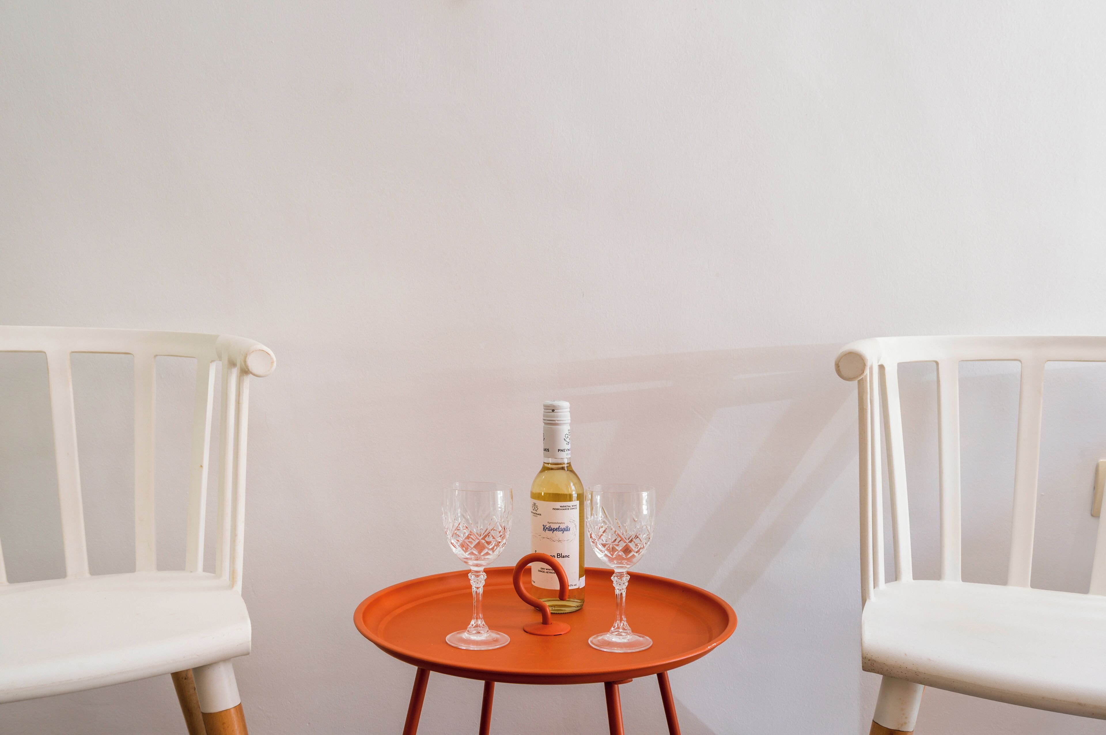 A white chair, a small orange table, a bottle of wine, and two wine glasses.