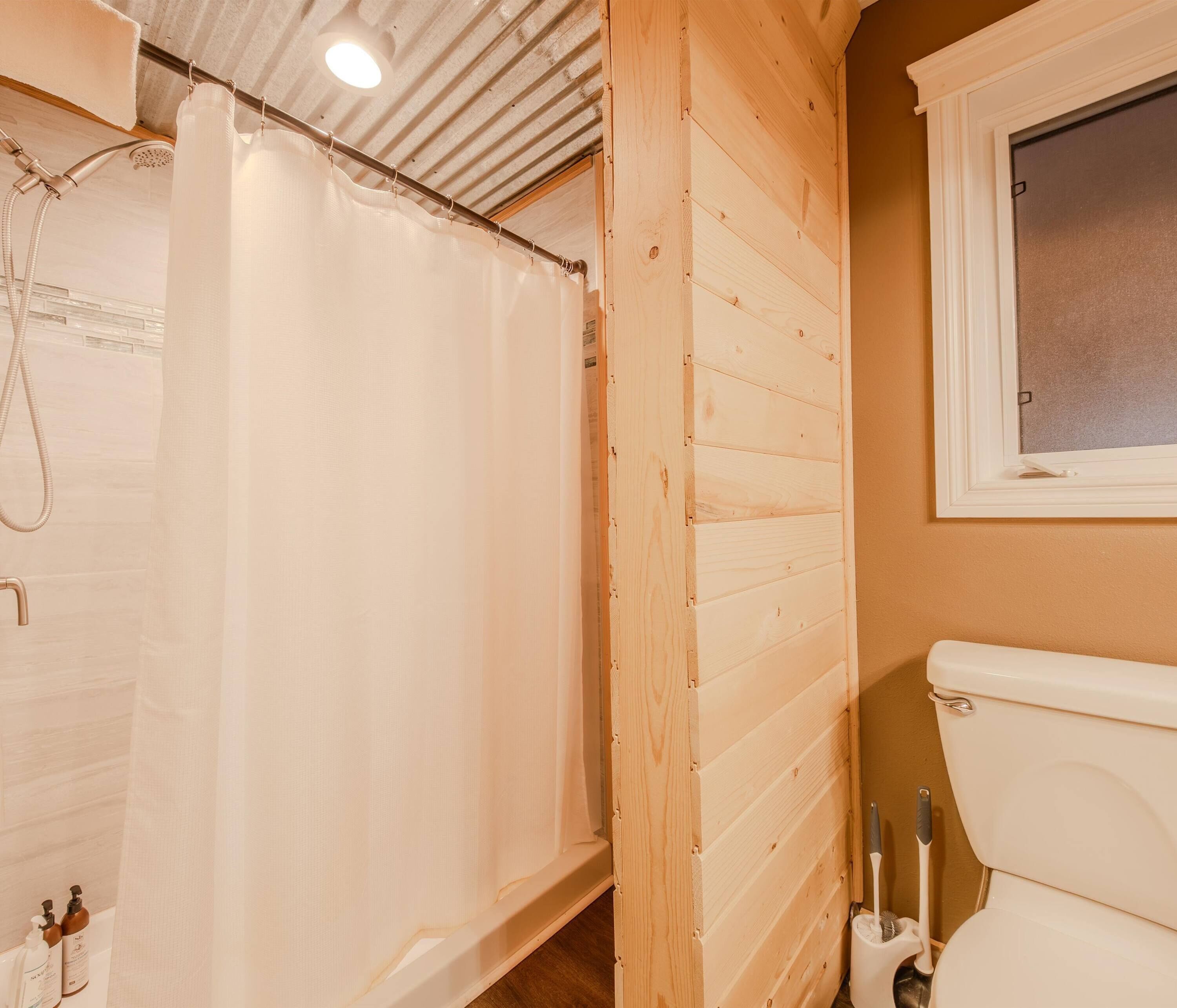 A bathroom with a shower curtain, wooden panel walls, a toilet, and a window.