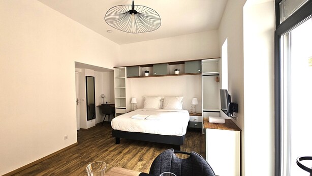 Une chambre d’hôtel moderne, dotée d’un grand lit, d’un bureau, d’une chaise, d’une télévision et d’un coin repas avec une table dressée pour deux personnes.