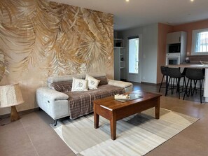 Un salon comprenant un canapé, une table basse et un mur orné d’un motif de feuilles. Il y a également un coin cuisine avec une table à manger et des chaises.
