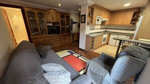 A living room with a grey sofa, a grey armchair, and a patterned rug. There is a wooden cabinet with glass doors and a microwave above the counter. - Apartment for 2 in the center of Nueva (Nueva de Llanes)