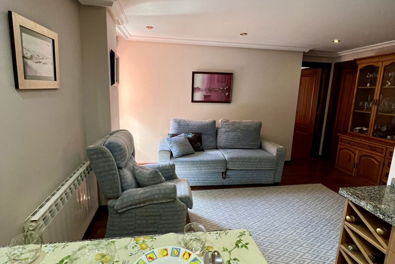 A living room with a grey sofa, a grey armchair, a patterned rug, and a dining table with a white tablecloth.