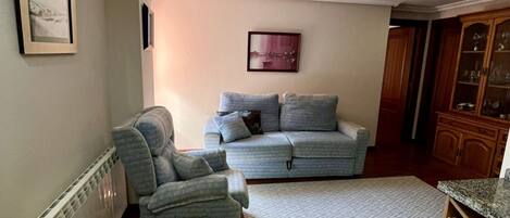 A living room with a grey sofa, a grey armchair, a patterned rug, and a dining table with a white tablecloth.