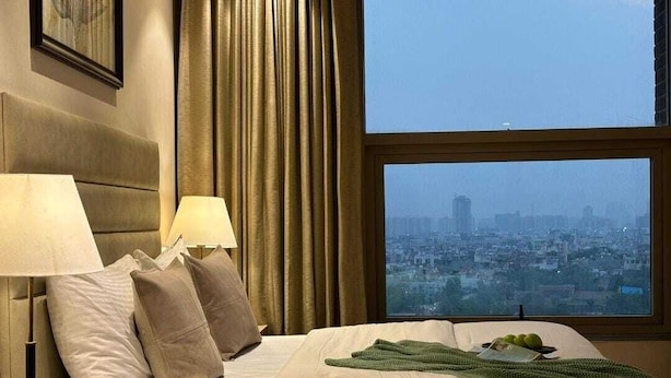 Una habitación de hotel con cama, almohadas, lámpara, ventana y vista a la ciudad, además de una manta verde.