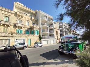 Une rue où l’on aperçoit un autobus vert portant l’inscription « SPINOLA » et une voiture blanche garée sur le bord.
