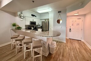 A kitchen with a central island, bar stools, a microwave, refrigerator, and a hanging fish decoration. - Master on Gulf, Lagoon Pool & Beach Views w/ Free Daily Activities! (Panama City Beach)