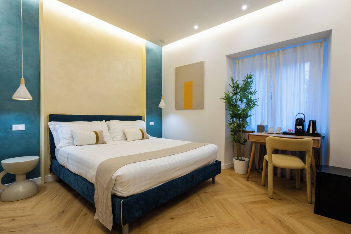 A bedroom with a bed, bedside table, lamp, desk, chair, and a potted plant.