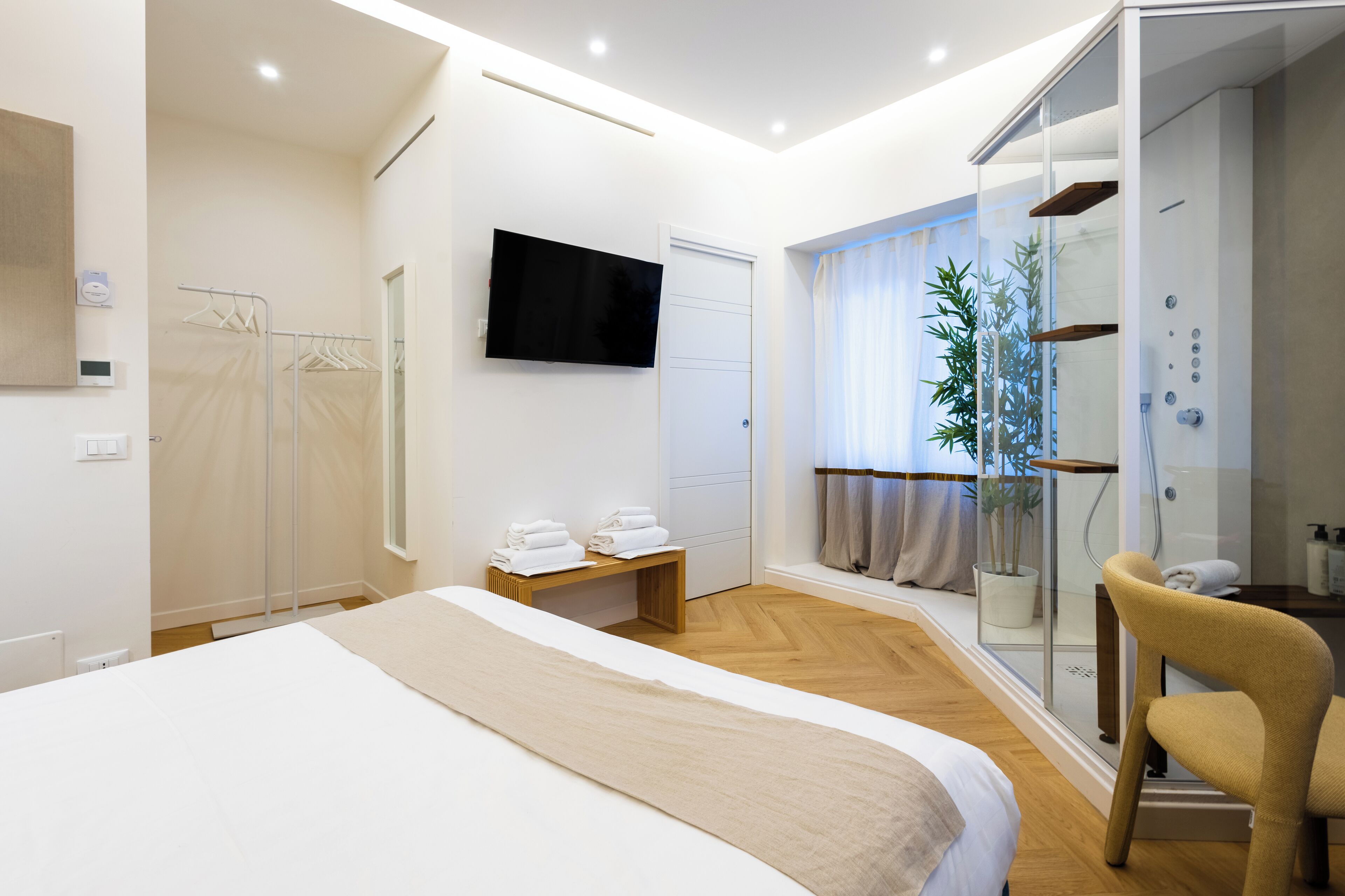A modern hotel room with a large bed, a TV mounted on the wall, a shower area with a glass door, and a small seating area with towels.