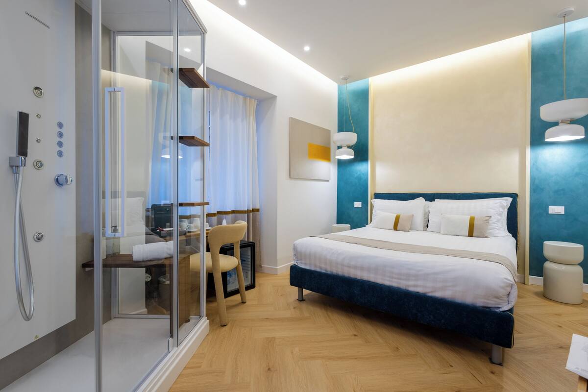 A modern hotel room with a glass shower enclosure, a bed with white and blue bedding, and a wooden floor.