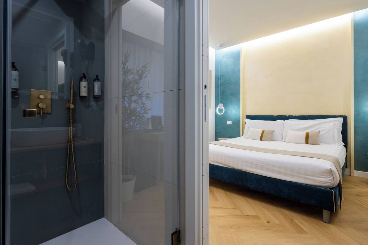 A modern bathroom with a glass-enclosed shower, a bed with a blue headboard, and a bedside table.