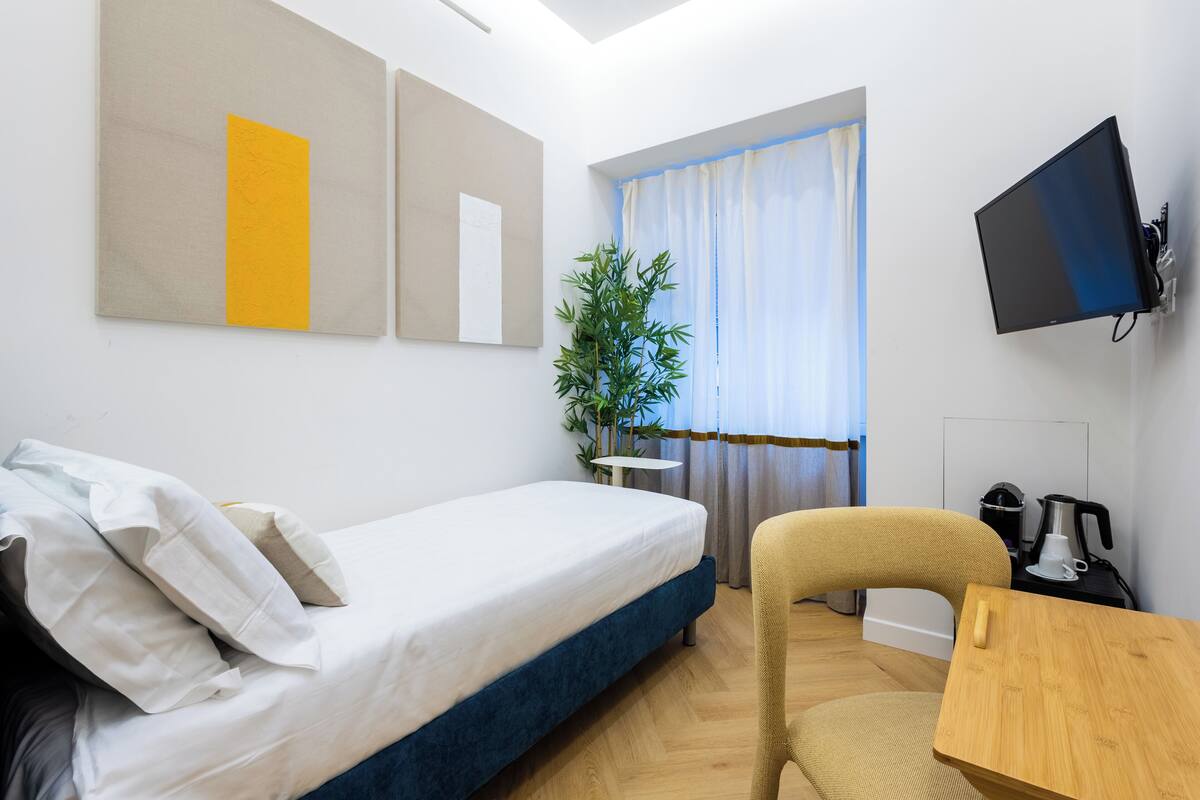 A hotel room with a bed, a chair, a small table, a TV, and a window with curtains.