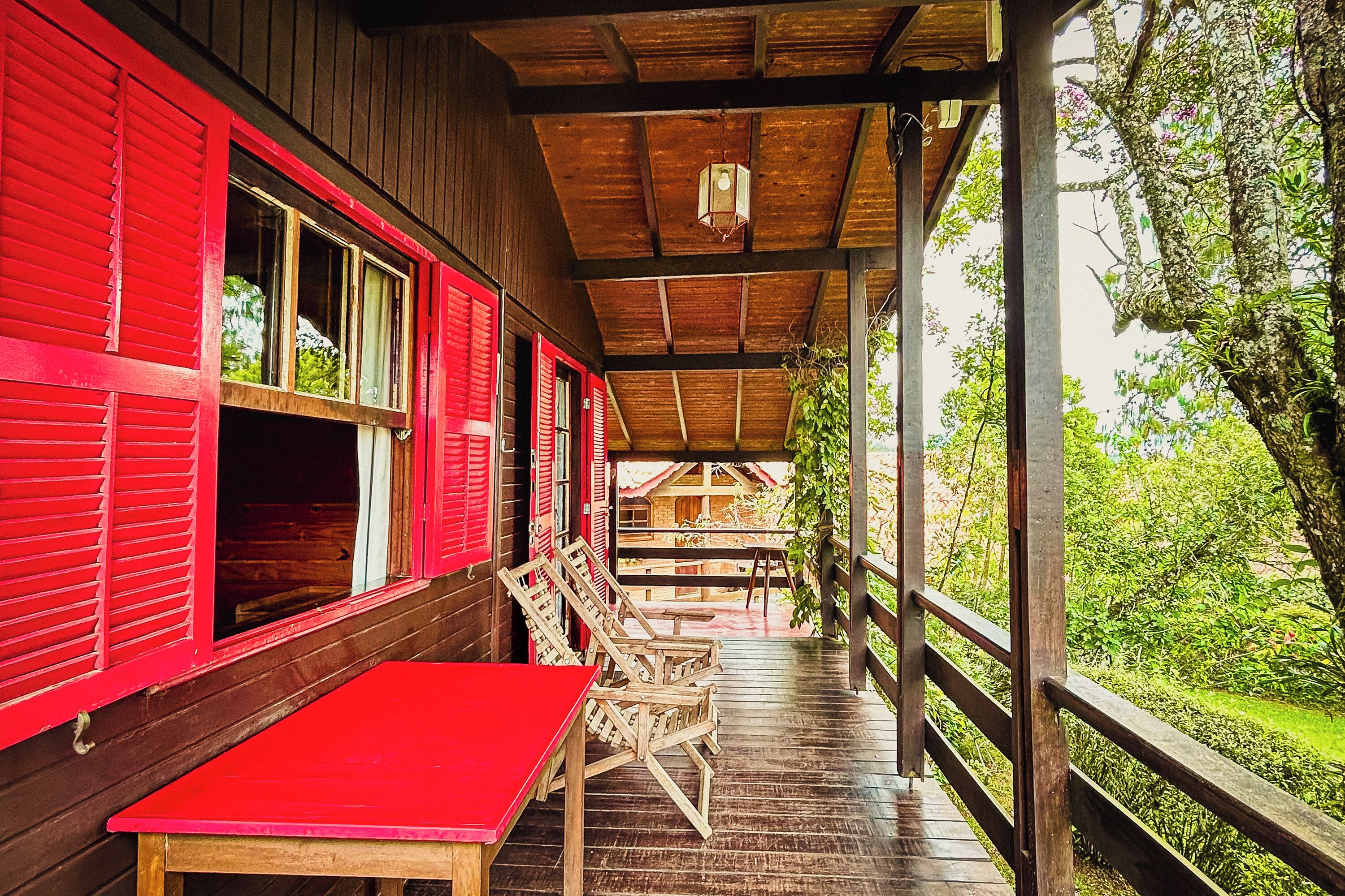 A wooden building with red shutters, a covered porch with wooden chairs, and a view of greenery.