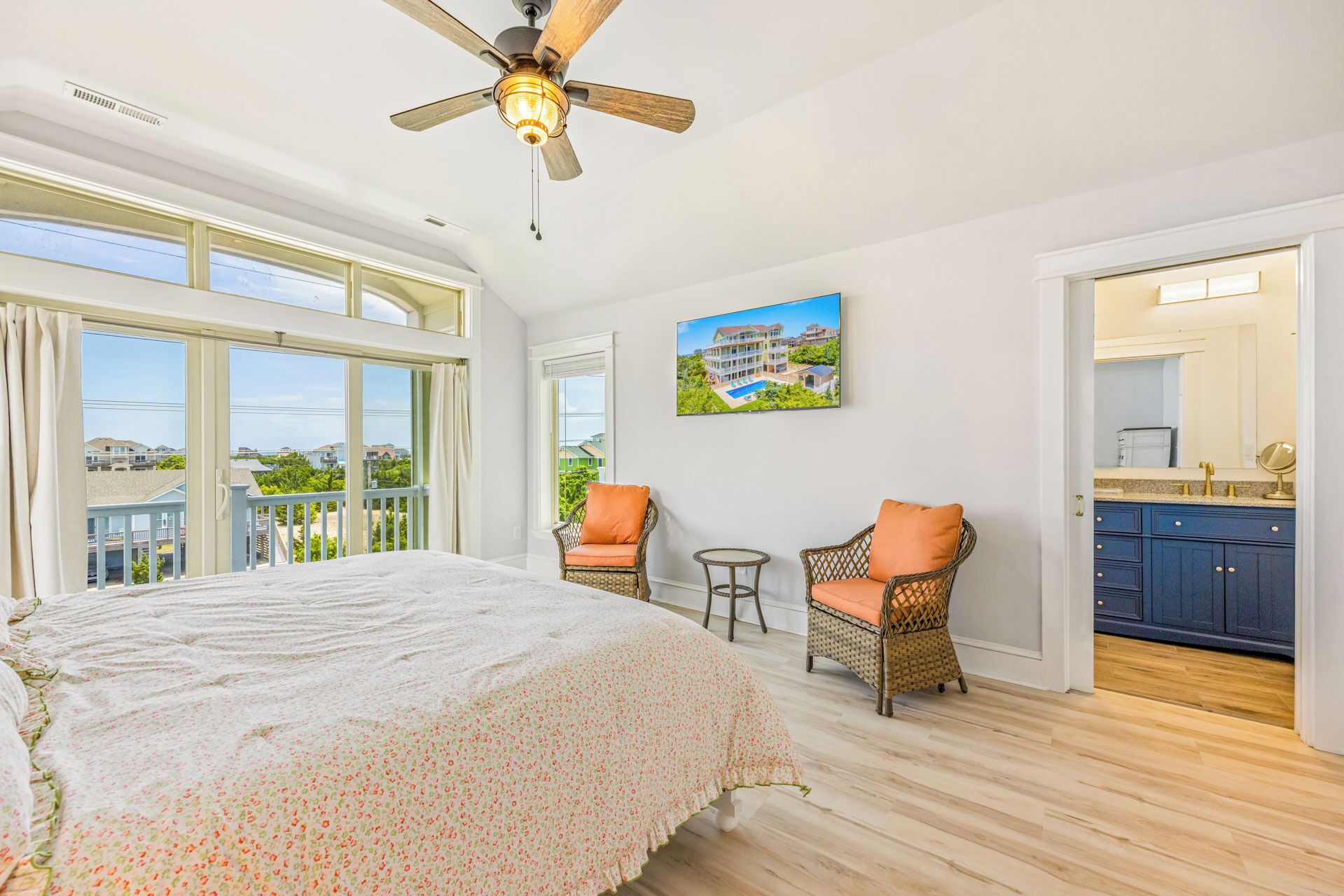 A bedroom with a large bed, a ceiling fan, a view of the outside, a small table, two chairs, and a kitchen area.