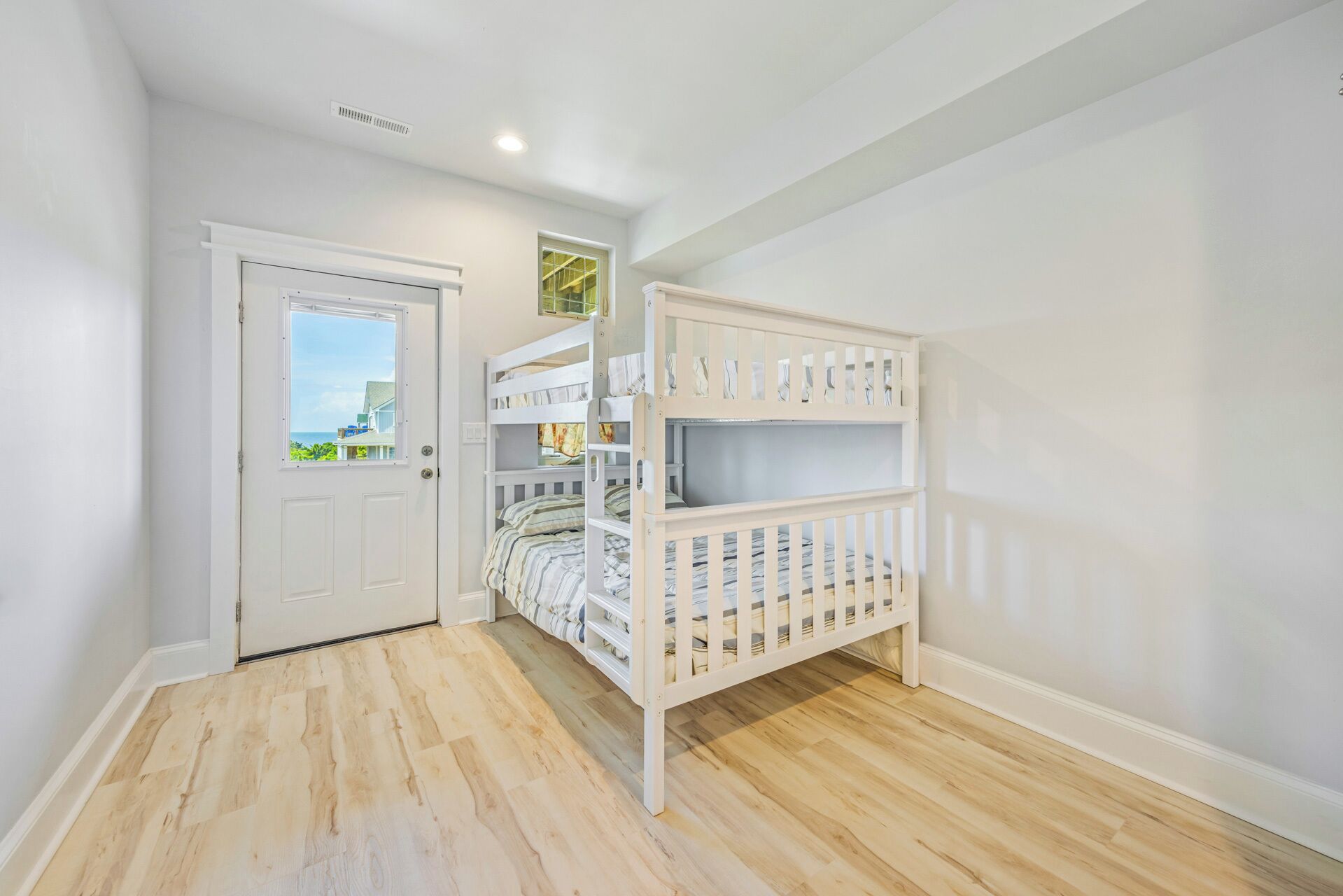 A room with a white door, a window, a bunk bed, and wooden flooring.