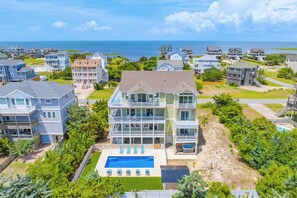 A multi-story residential building with a swimming pool, surrounded by other houses, and a view of the ocean. - New Soundside Dog Friendly Avon Retreat with Pool, Hot Tub, Game Room, Theater (Salvo)