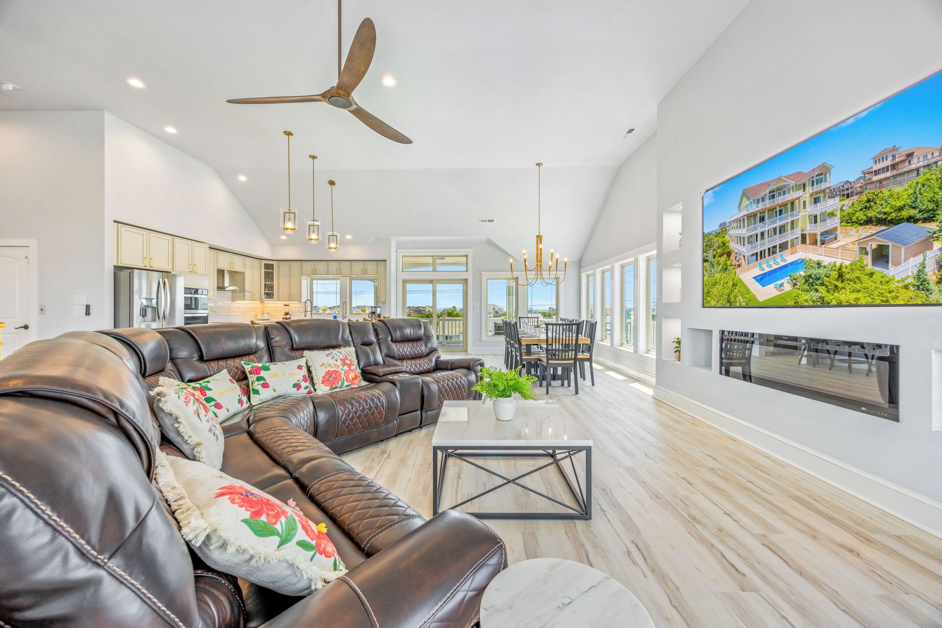 A spacious living room with a leather sofa, a dining area, a kitchen, and a large TV displaying a residential property.